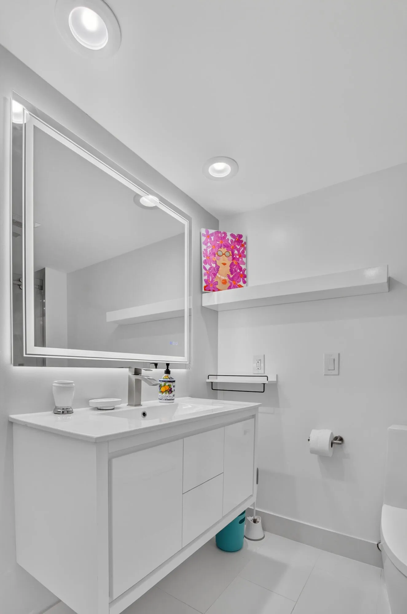 Recessed ceiling lighting illuminates a white vanity with integrated sink, a large backlit mirror, and a wall-mounted floating shelf