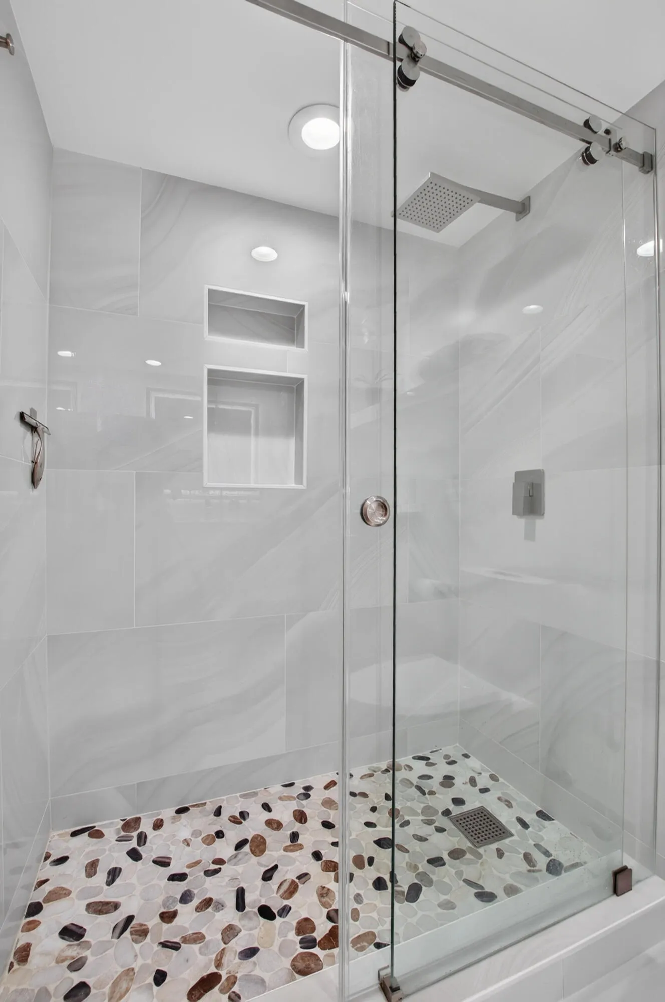 Glass-enclosed shower with a multi-tone pebble floor, built-in recessed shelving, a wall-mounted rain shower head, and an overhead recessed light