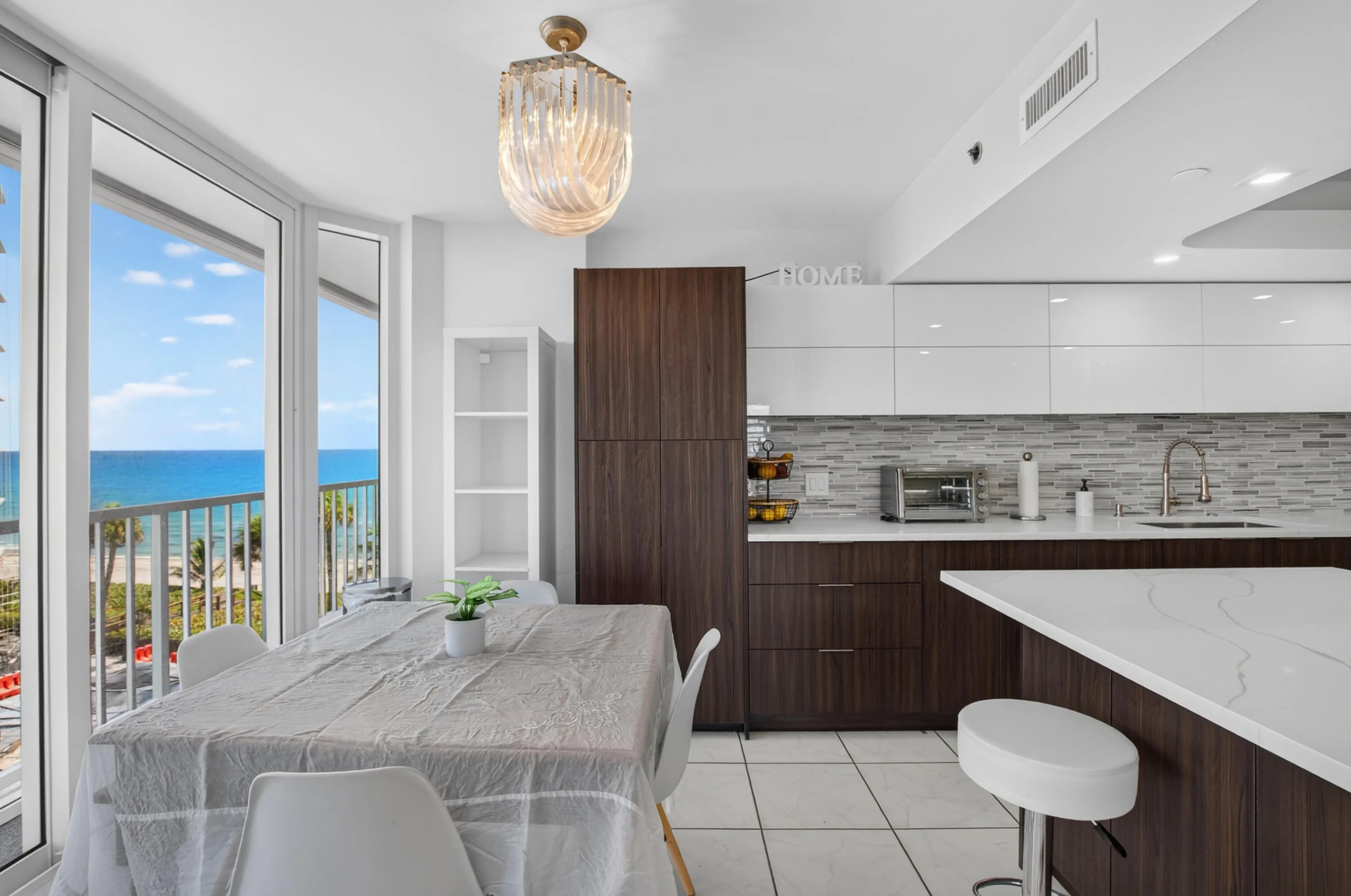 Floor-to-ceiling balcony doors, contemporary kitchen cabinetry, light-toned tile flooring, island with countertop seating, and a mosaic tile backsplash