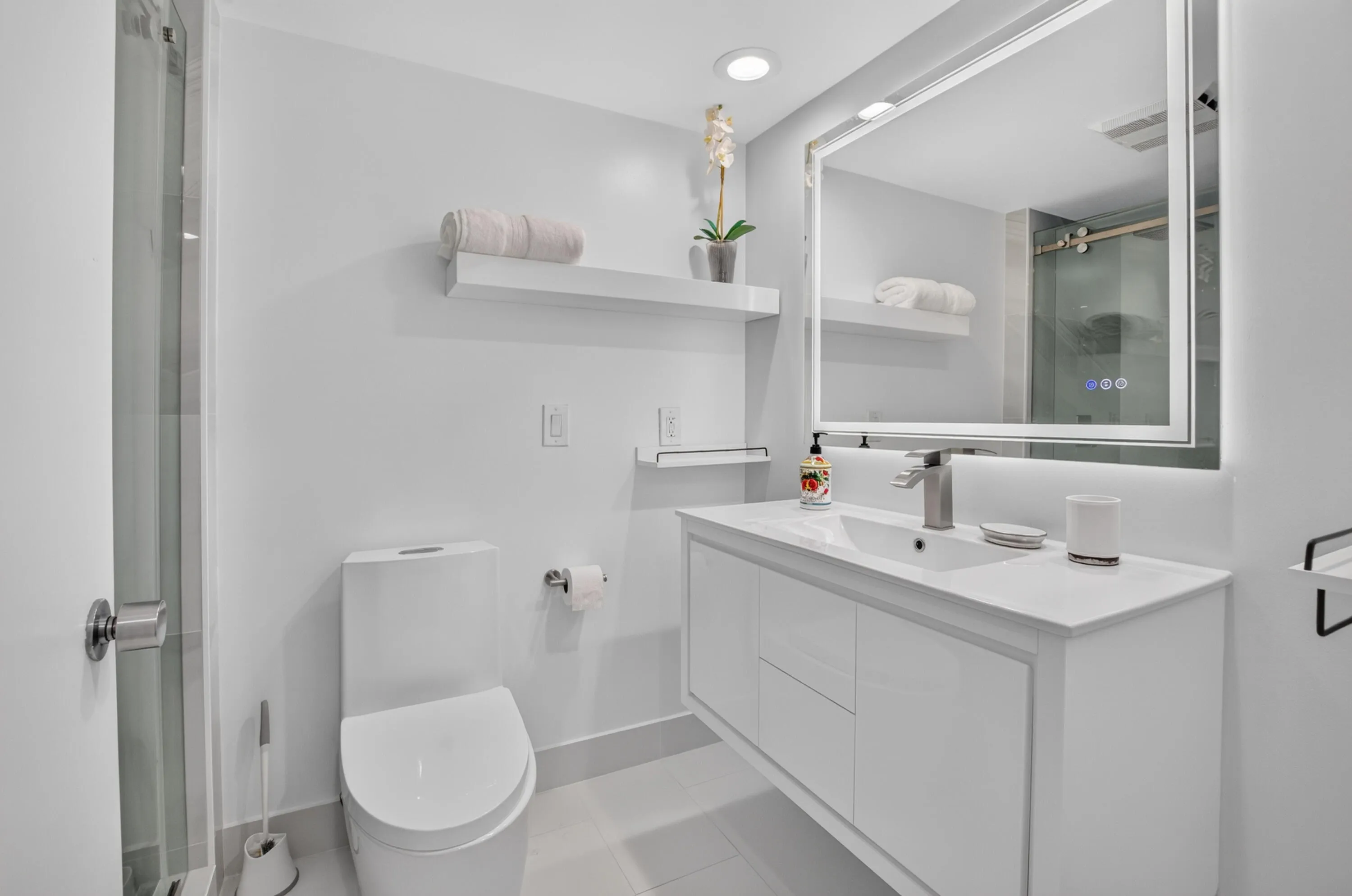 Floating vanity with integrated sink, backlit wall-mounted mirror, recessed lighting, and a frameless glass shower enclosure