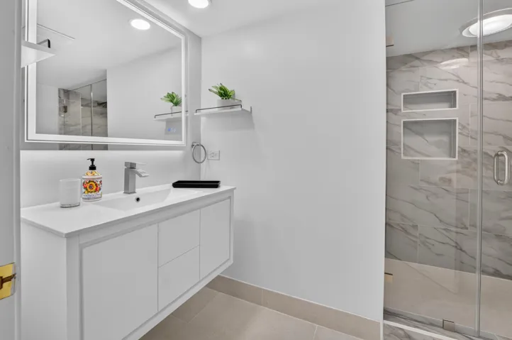 Floating vanity with integrated sink, illuminated mirror, walk-in shower with glass enclosure, marble-patterned tile, and recessed shower niches