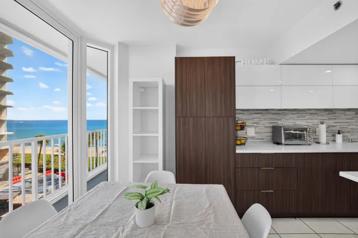 Expansive glass doors opening to a balcony, modern kitchen cabinetry with wood-finish and white high-gloss finishes, contemporary linear backsplash, integrated shelving, and a prominent ocean backdrop