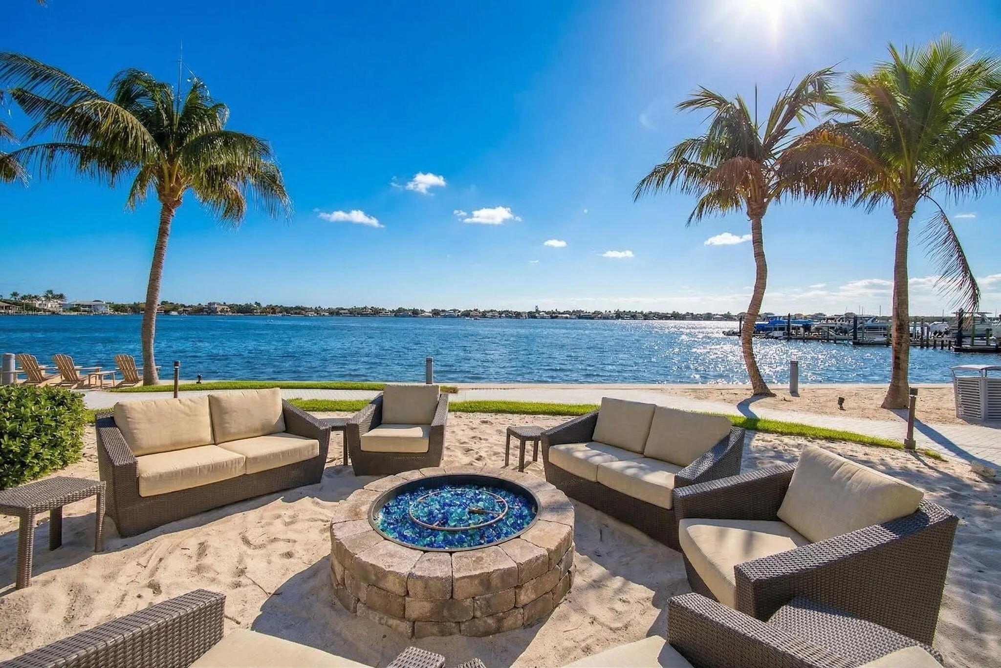 Waterfront setting featuring a sandy lounging area, stone fire pit, and mature palm trees