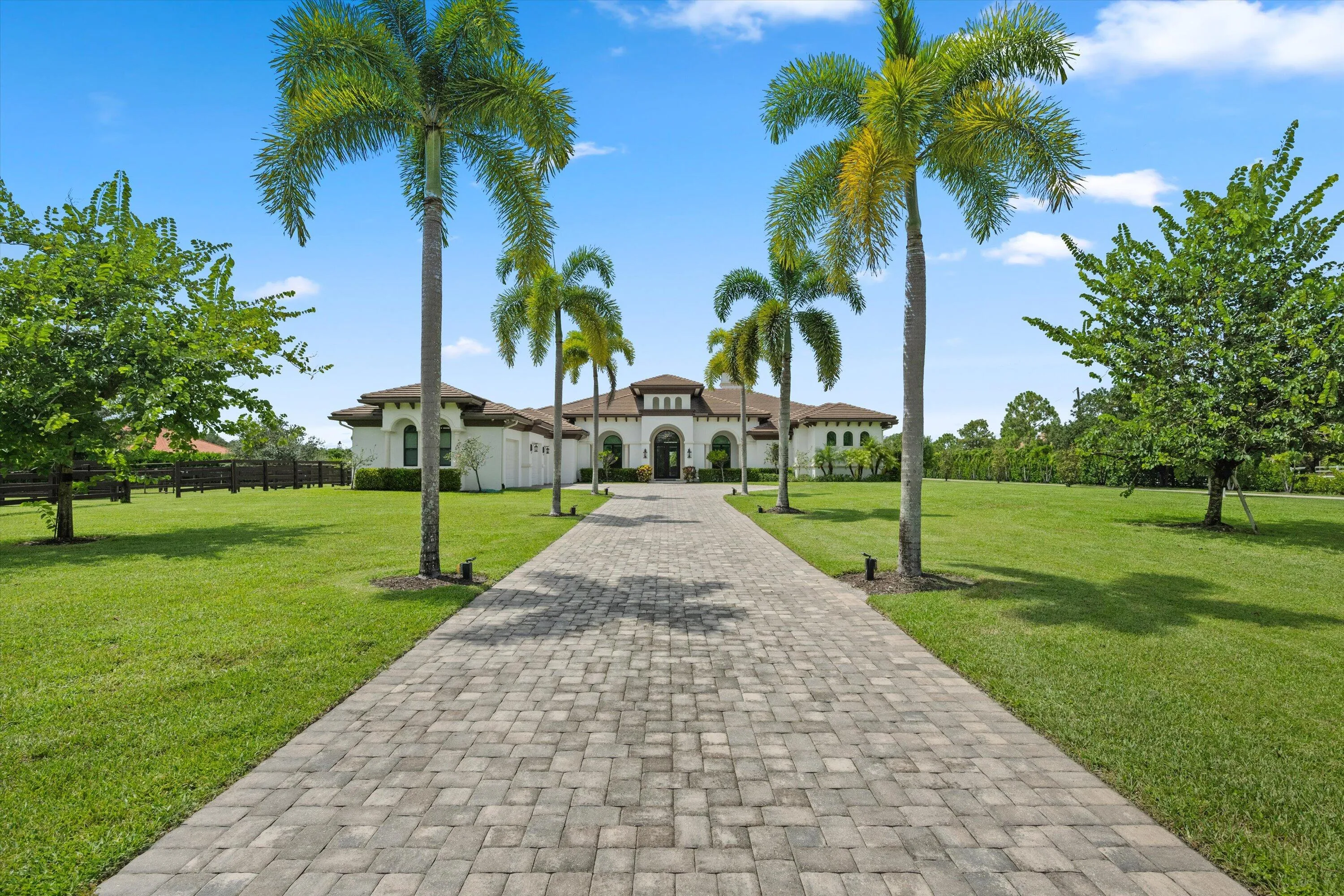 Paver driveway framed by mature palm trees, expansive green lawn, and a stucco exterior with arched windows