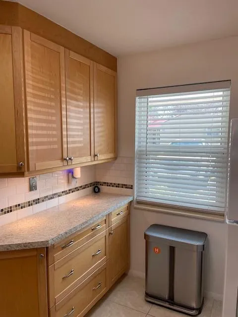 Natural wood-finish cabinetry with silver-tone hardware, light-toned countertop, subway tile backsplash with decorative mosaic accent, and tiled flooring