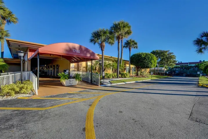 Covered entry with address signage, flanked by a paved driveway and landscaped grounds featuring palm trees and shrubs