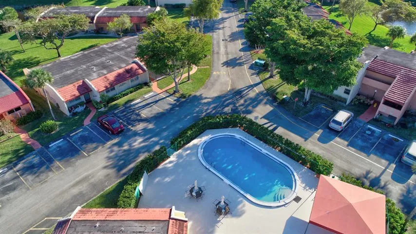 Oval-shaped outdoor swimming pool with surrounding hardscaping, mature perimeter landscaping, and adjacent parking areas
