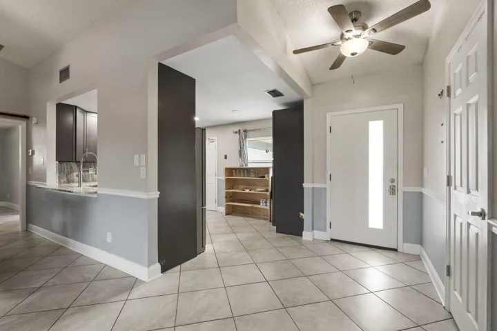 Tiled flooring throughout, modern entry door with vertical glass insert, ceiling fan with integrated lighting, two-tone wall treatment, and kitchen pass-through with countertop