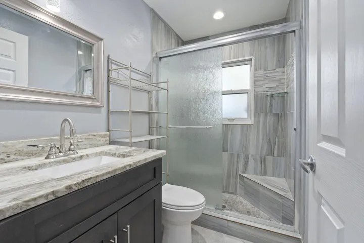 Granite-look countertop with an undermount sink, dark wood-finish vanity, framed wall mirror, and a tiled shower with a built-in bench