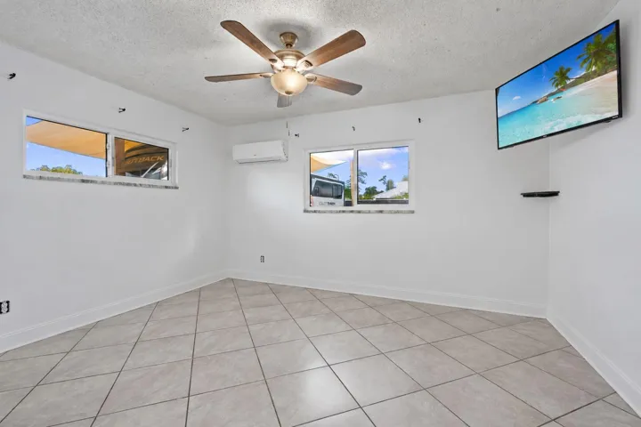 Ceiling fan with light fixture, white walls, multiple windows, wall-mounted air conditioning unit, and tile flooring
