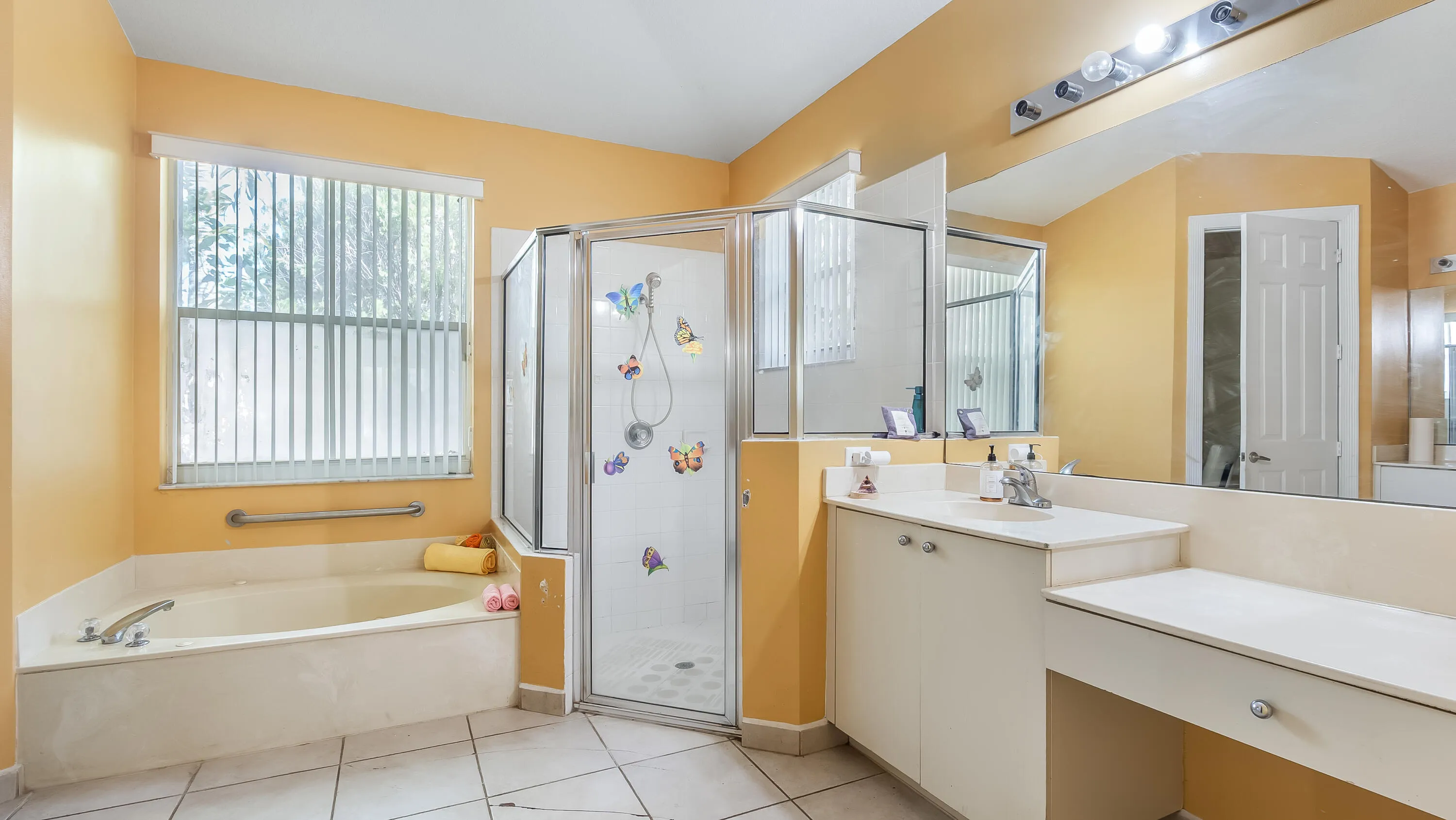 Corner soaking tub with grab bar, glass-enclosed shower, dual vanity with undermount sink, built-in makeup vanity, and tiled flooring
