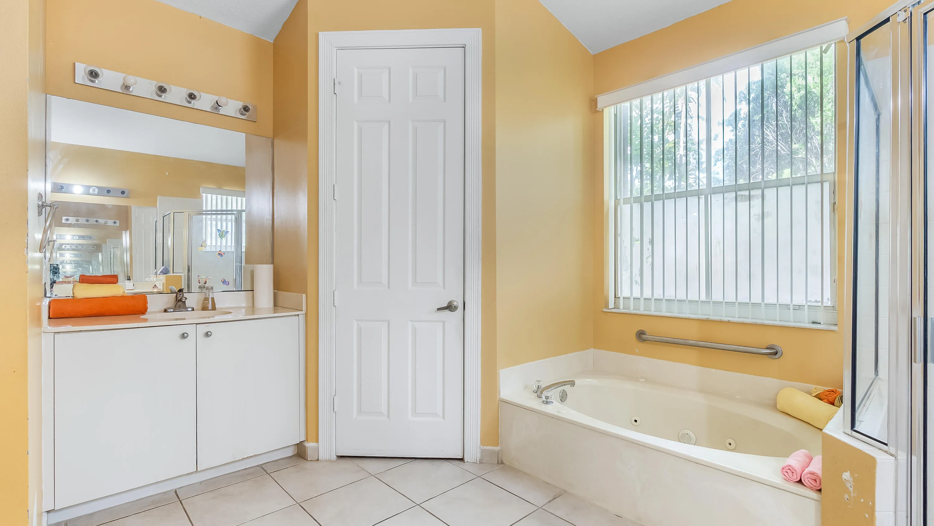 Built-in corner soaking tub with window, glass-enclosed shower, white vanity with integrated sink, large mirror, and tiled flooring