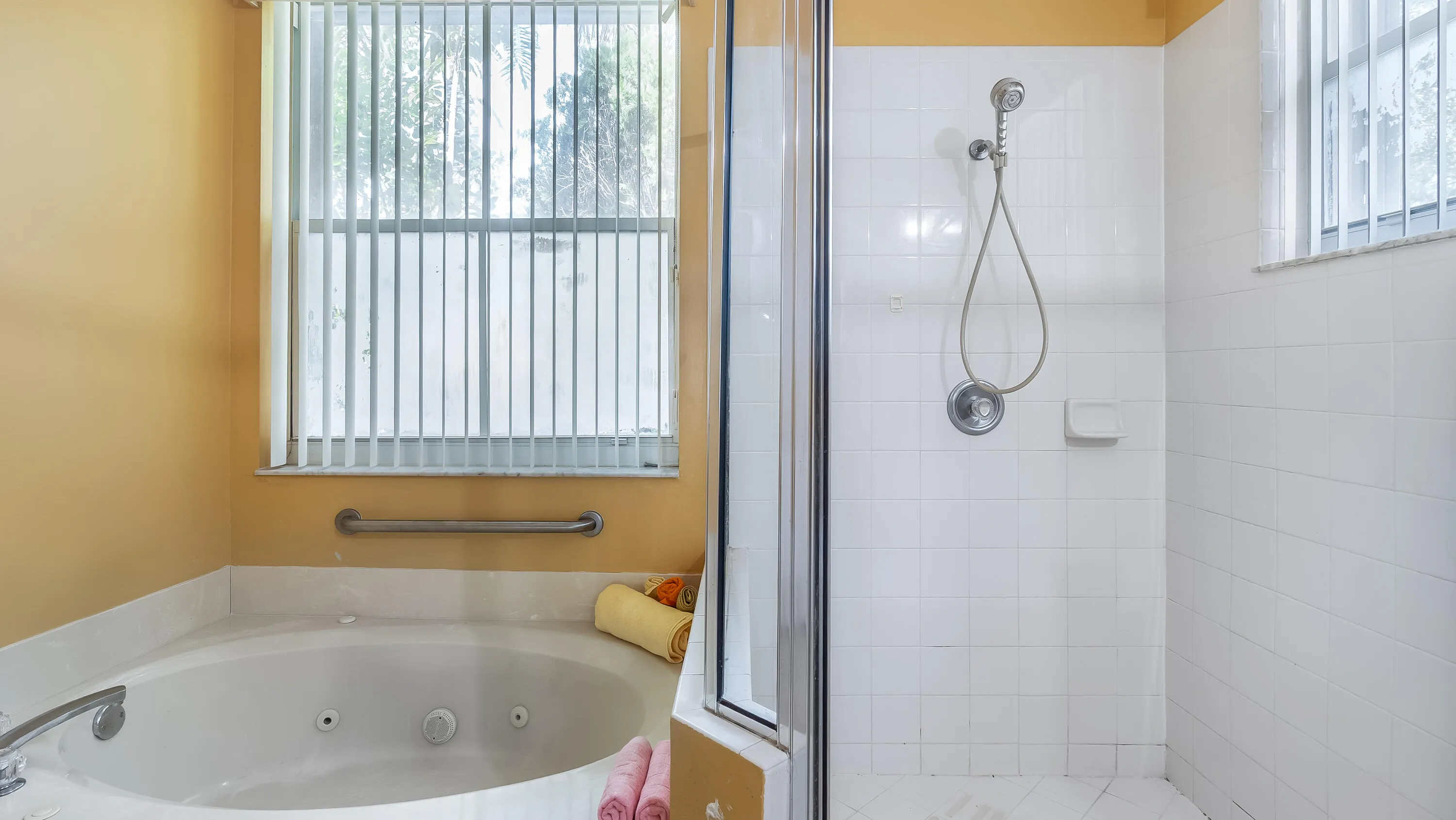 Built-in jetted tub with integrated grab bar, separate shower with tiled walls, and privacy window blinds
