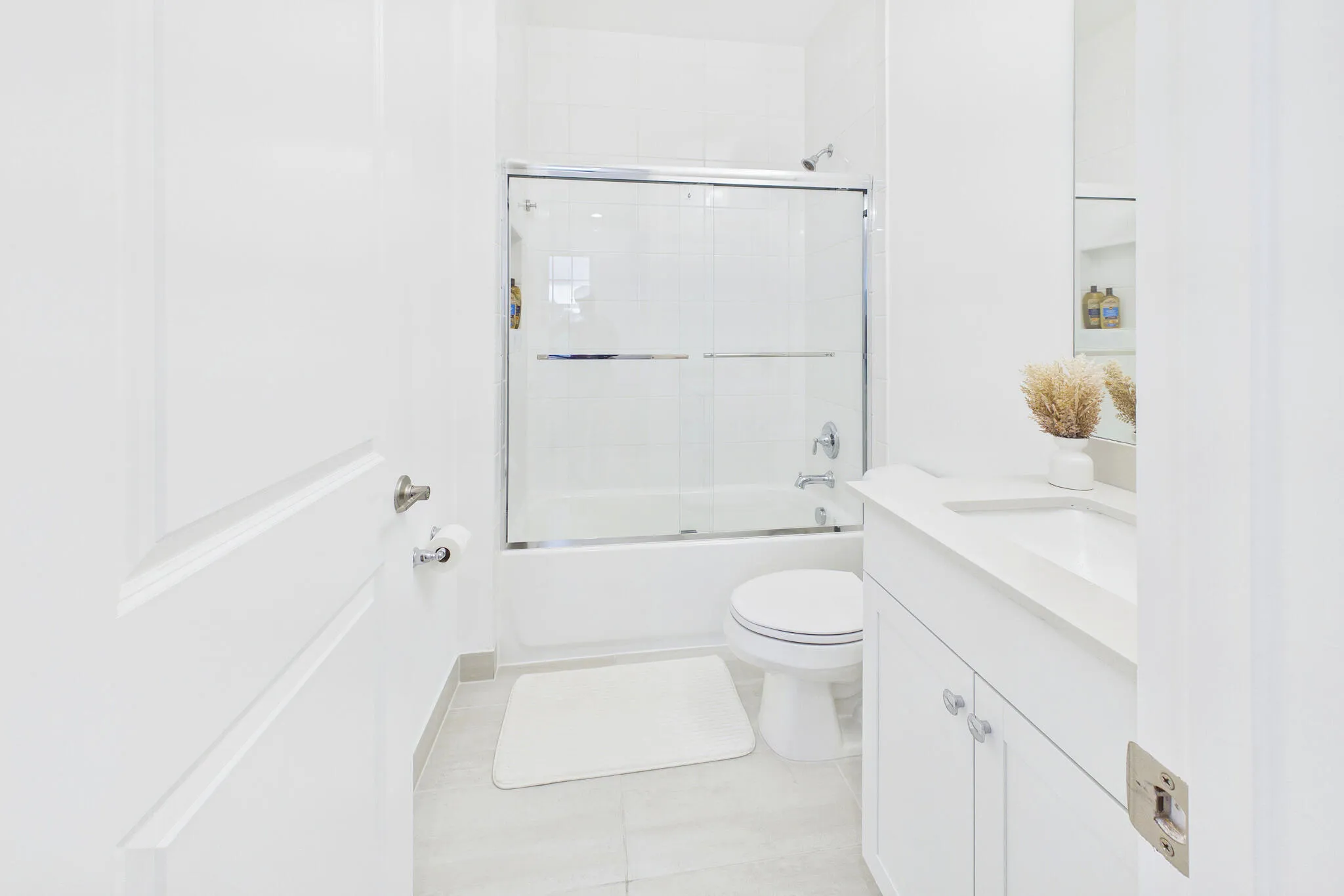 White bathroom with light tile flooring, a bathtub with a sliding glass door, a toilet, and a vanity with an integrated sink