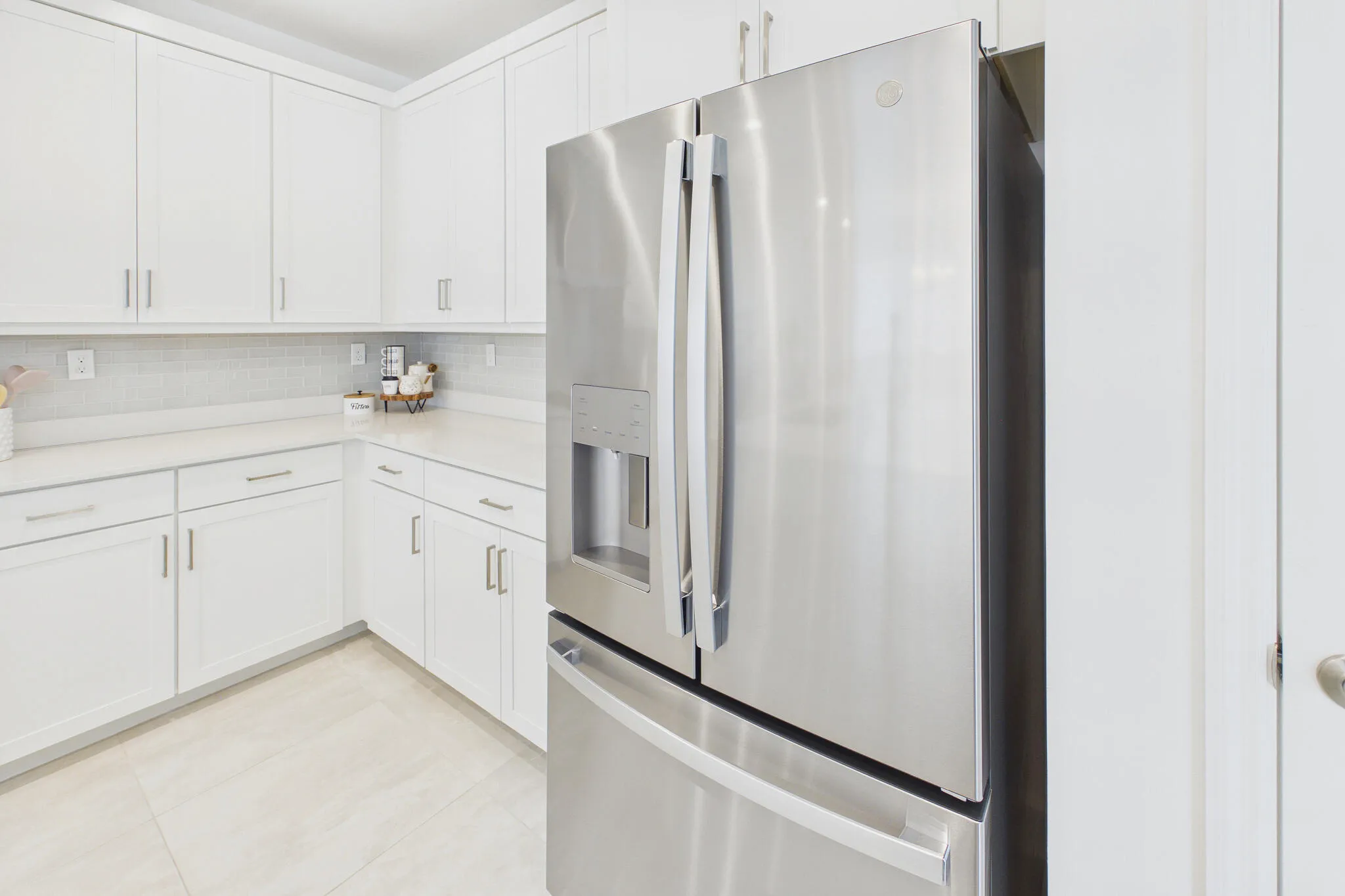 Stainless steel French door refrigerator, white shaker-style cabinetry, light-toned countertops, tile backsplash, and light-toned floor tile