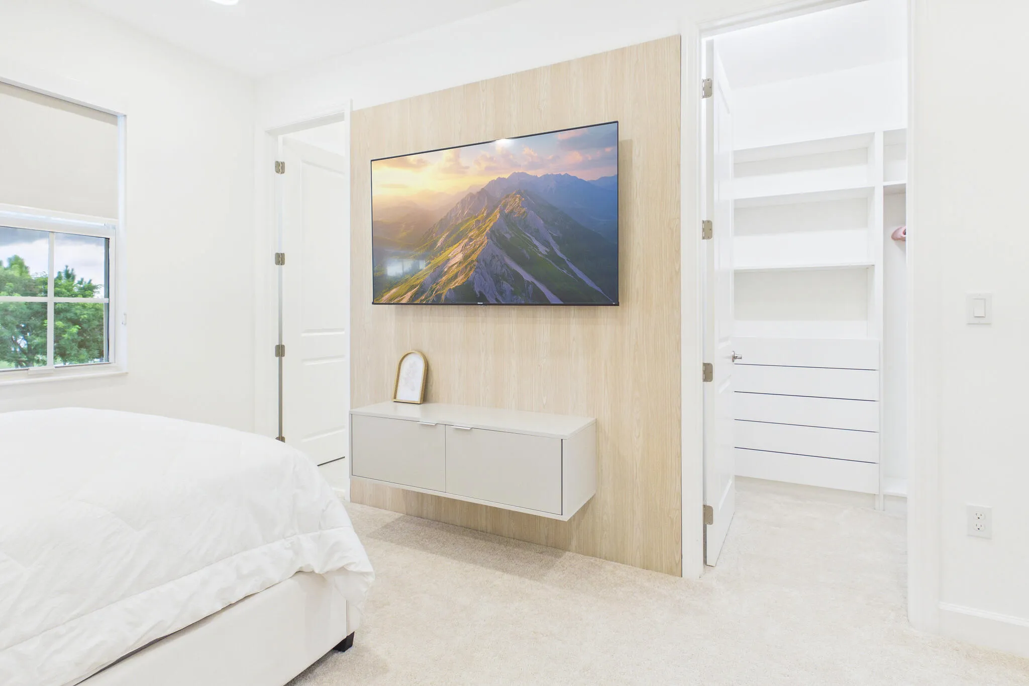 Built-in floating media console, wood-finish accent wall, window with roller shade, and an organized closet system featuring shelving and drawers
