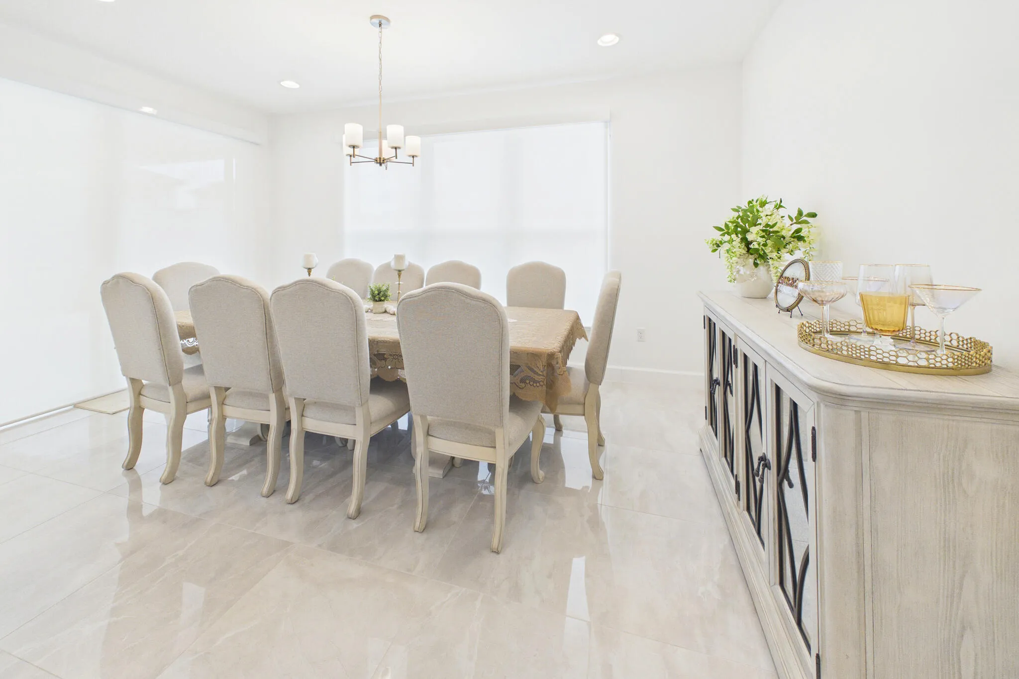Recessed lighting, chandelier, and large window with roller shades illuminate a room featuring light-toned tile flooring and a built-in buffet cabinet