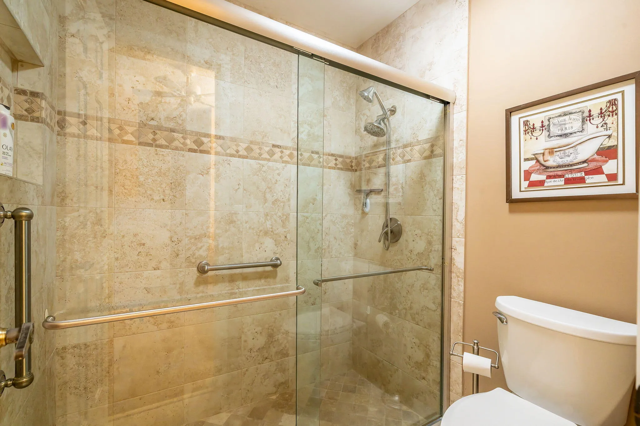 Glass-enclosed shower with stone tile surround, decorative mosaic tile band, and grab bars