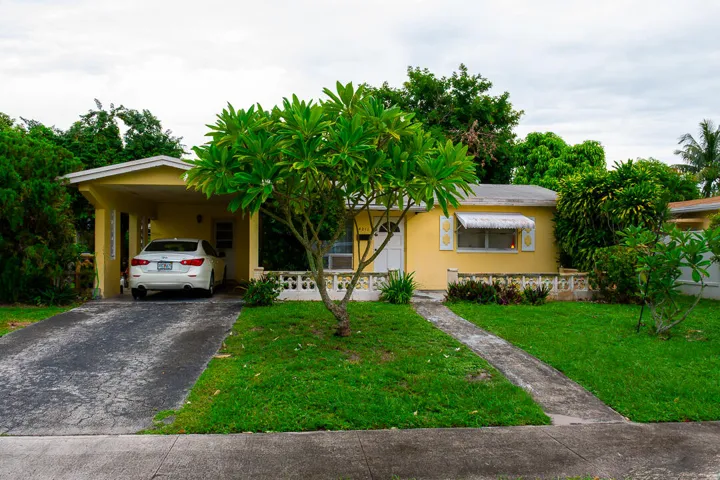 Single-story exterior with a yellow facade, covered carport, front lawn, and mature trees