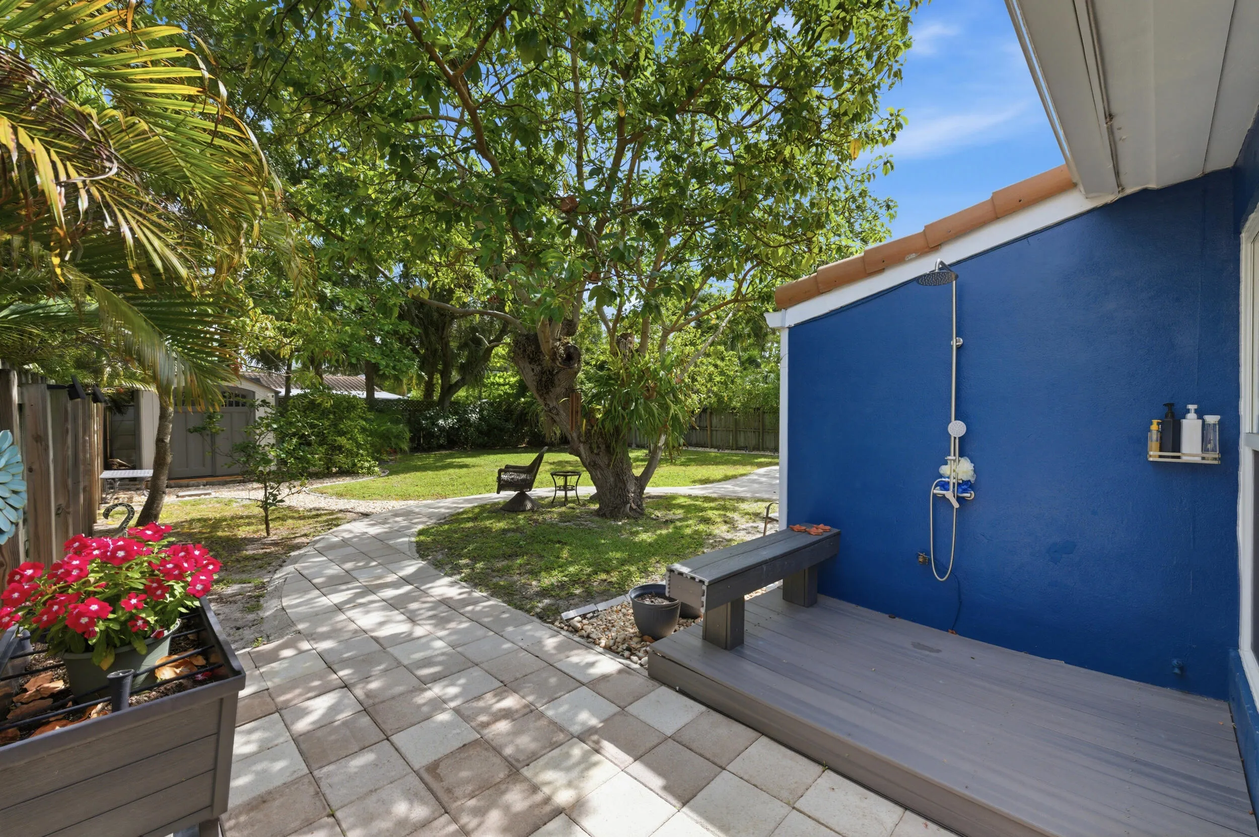 Paved pathway leading to a landscaped yard with mature trees, a composite deck, and an exterior shower fixture