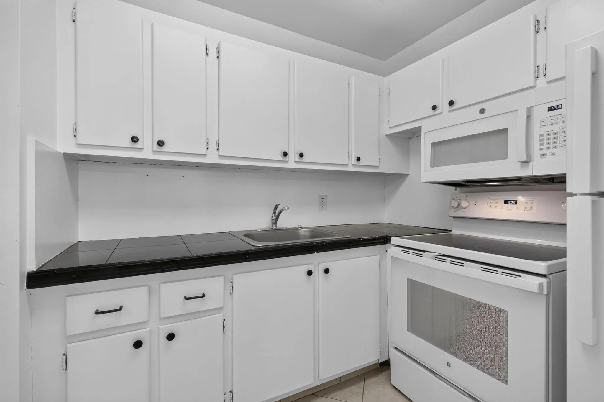 White cabinetry with black hardware, tile countertops, stainless steel sink, built-in microwave, and electric range