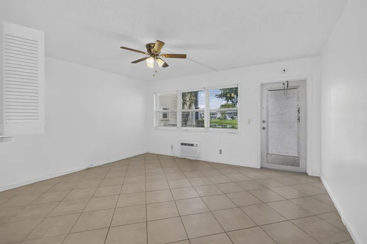 Tile flooring, ceiling fan with integrated lighting, multiple windows with blinds, a wall-mounted air conditioning unit, and an exterior door with a screen panel