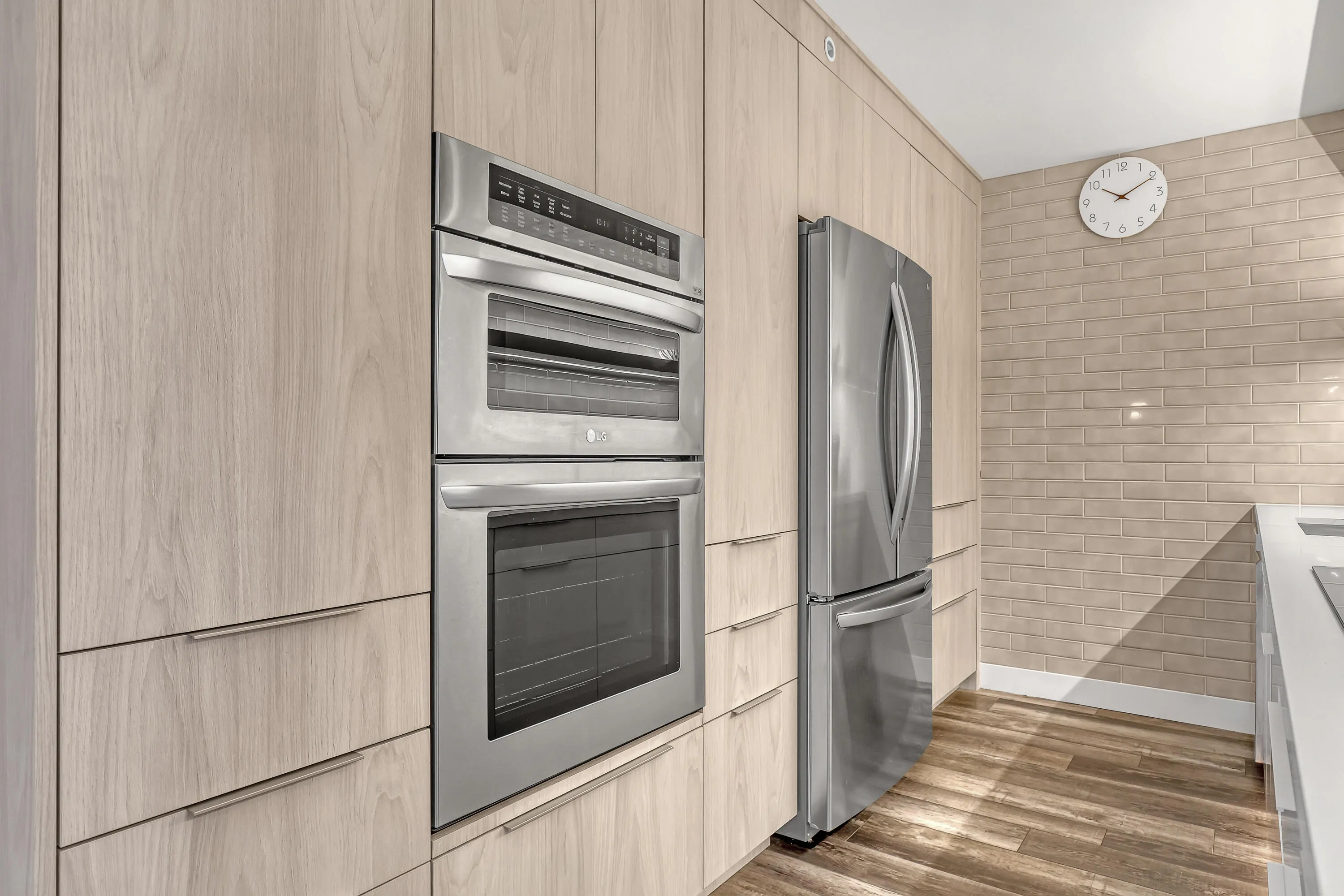 Integrated stainless steel double oven, stainless steel refrigerator, light wood-finish cabinetry, wood-finish flooring, and subway tile backsplash