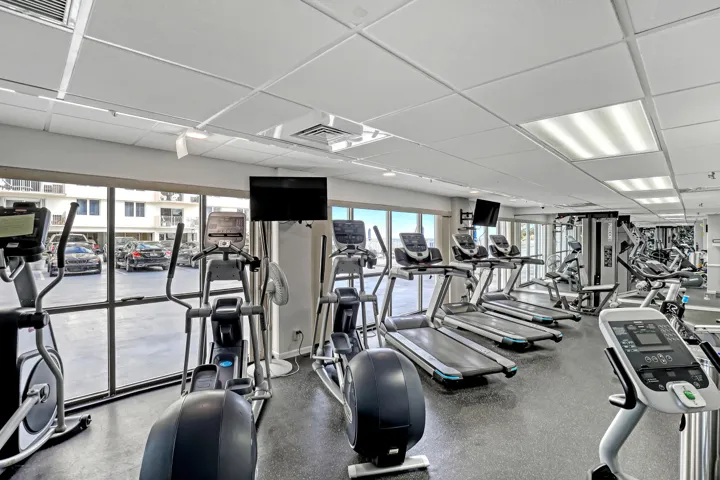 Fitness center with cardio equipment, strength training machines, wall-mounted televisions, large windows, and a grid ceiling with recessed lighting
