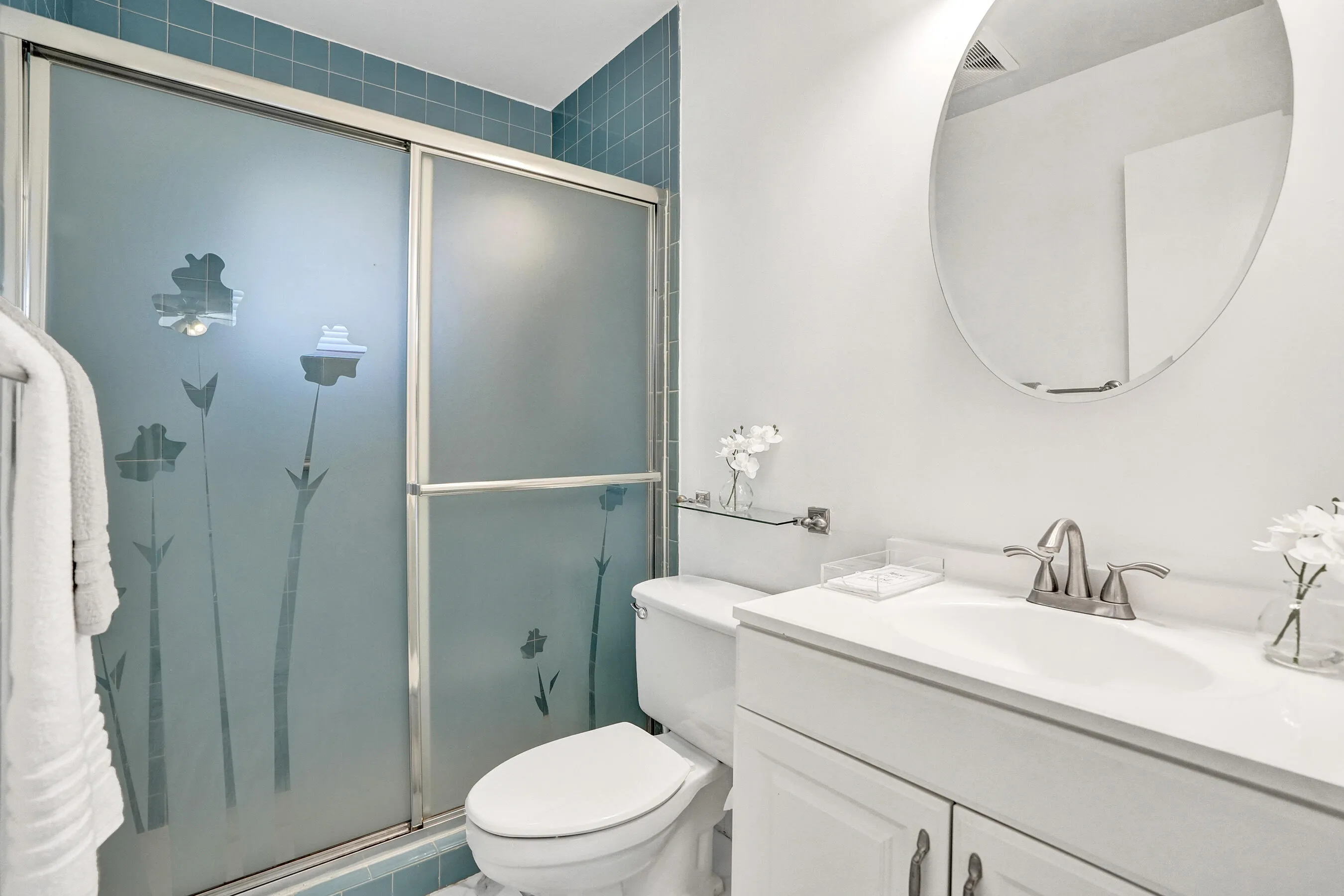 Shower with frosted glass enclosure, blue tile surround, white vanity with integrated sink, chrome-finish faucet, and an oval mirror