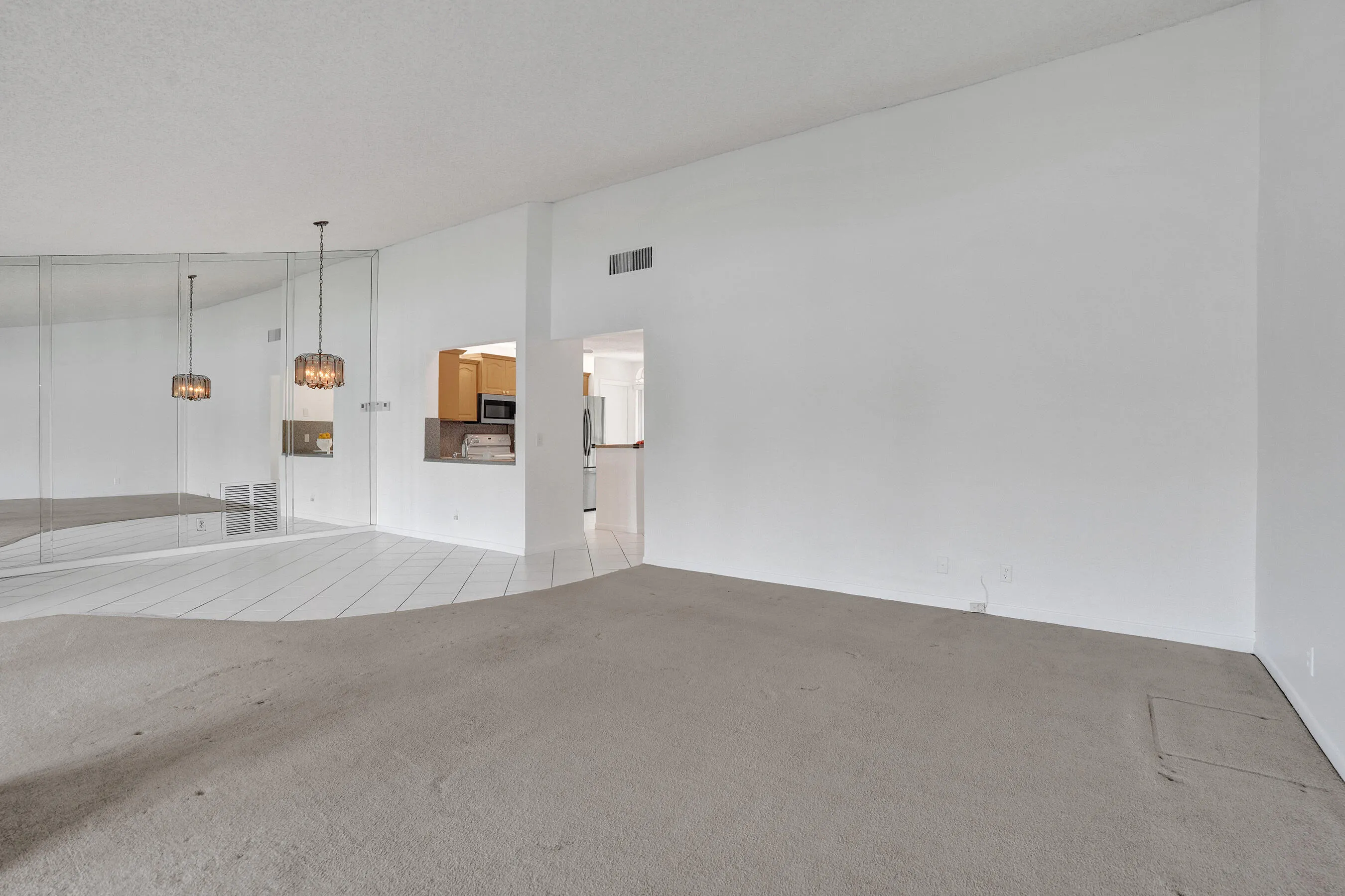Wall-to-wall mirrored accent wall, two suspended light fixtures, kitchen pass-through window with solid surface countertop, and mixed flooring with carpet and tile