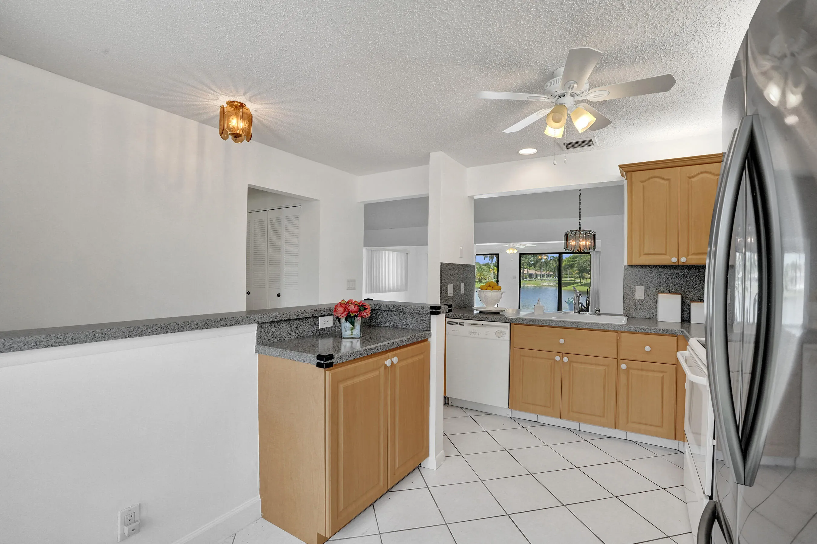 Kitchen featuring light wood cabinetry, white tile flooring, granite-finish countertops, and a stainless steel refrigerator