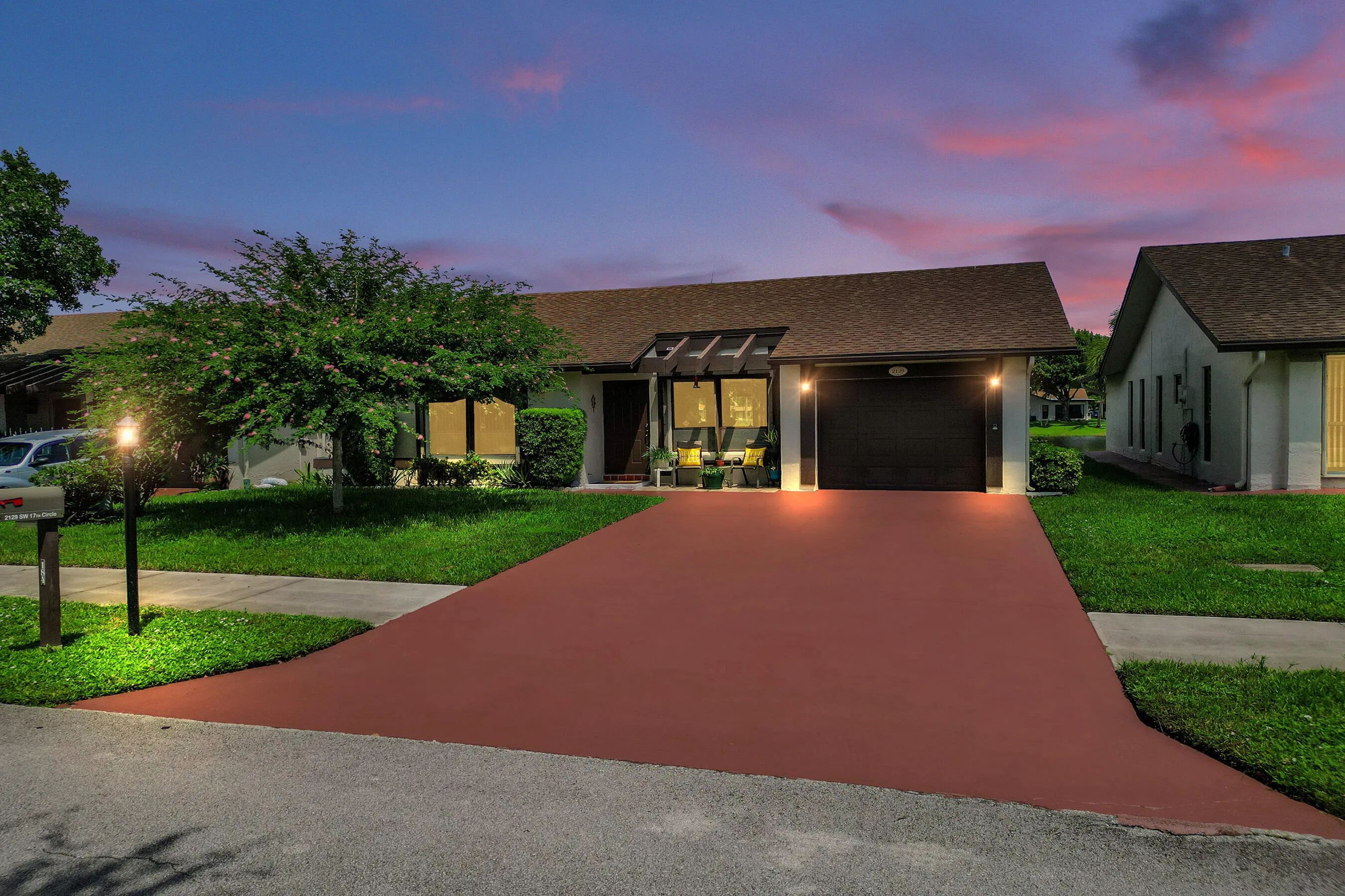 Red-hued driveway, single-car garage, front yard with mature tree, built-in exterior lighting, and sidewalk