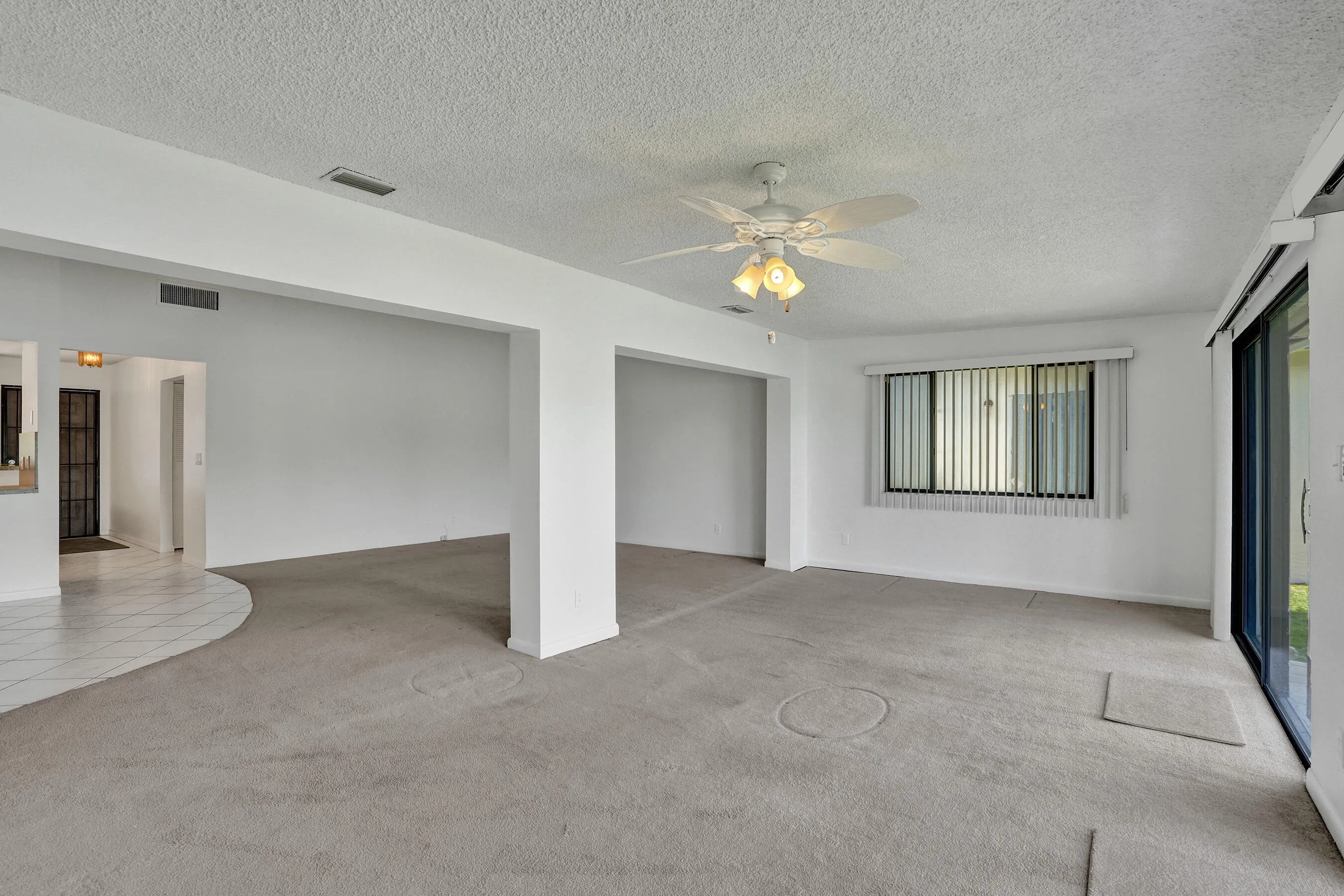Spacious common area featuring carpet flooring, a ceiling fan with integrated lighting, and a large window with blinds