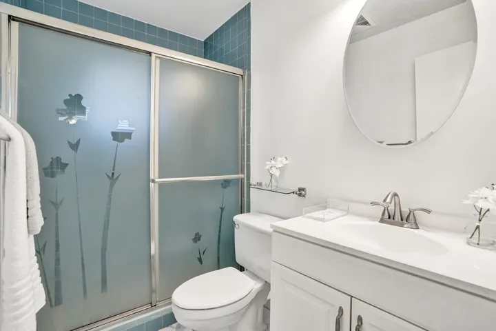 Shower with frosted glass enclosure, blue tile surround, white vanity with integrated sink, chrome-finish faucet, and an oval mirror
