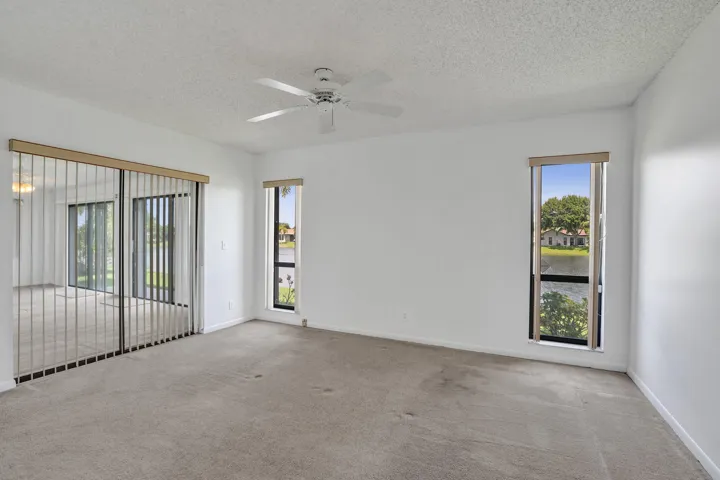 Expansive room with neutral carpeting, vertical blinds on sliding glass doors, two tall windows, and a ceiling fan