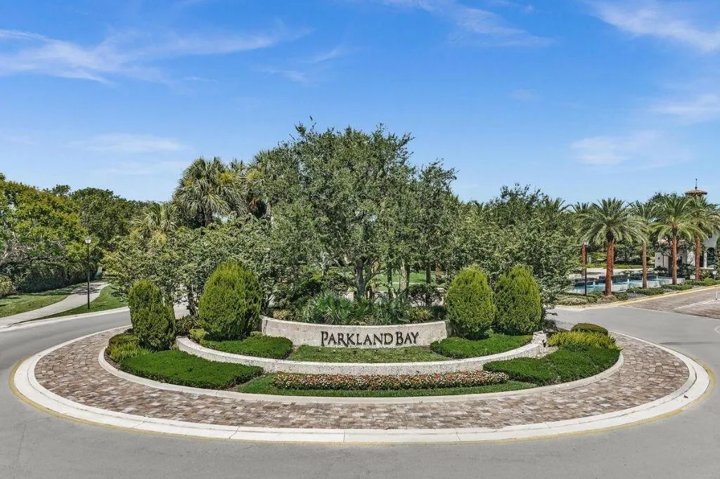 Roundabout entrance featuring a stone-clad "Parkland Bay" sign, lush landscaping, brick paver accents, and mature palm trees
