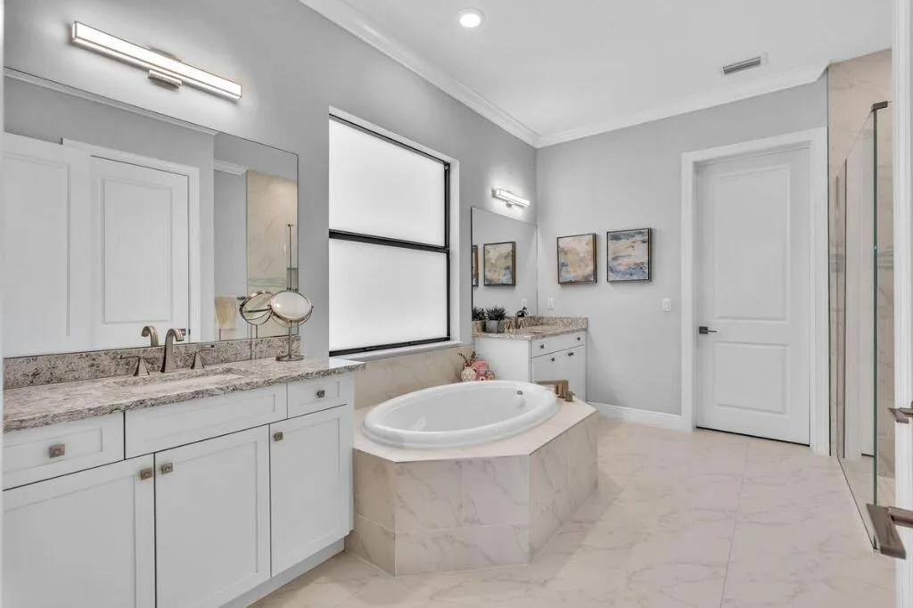 Light-filled bathroom featuring dual vanity stations with integrated sinks and granite countertops, a built-in soaking tub, a frameless glass shower enclosure, and large format tile flooring