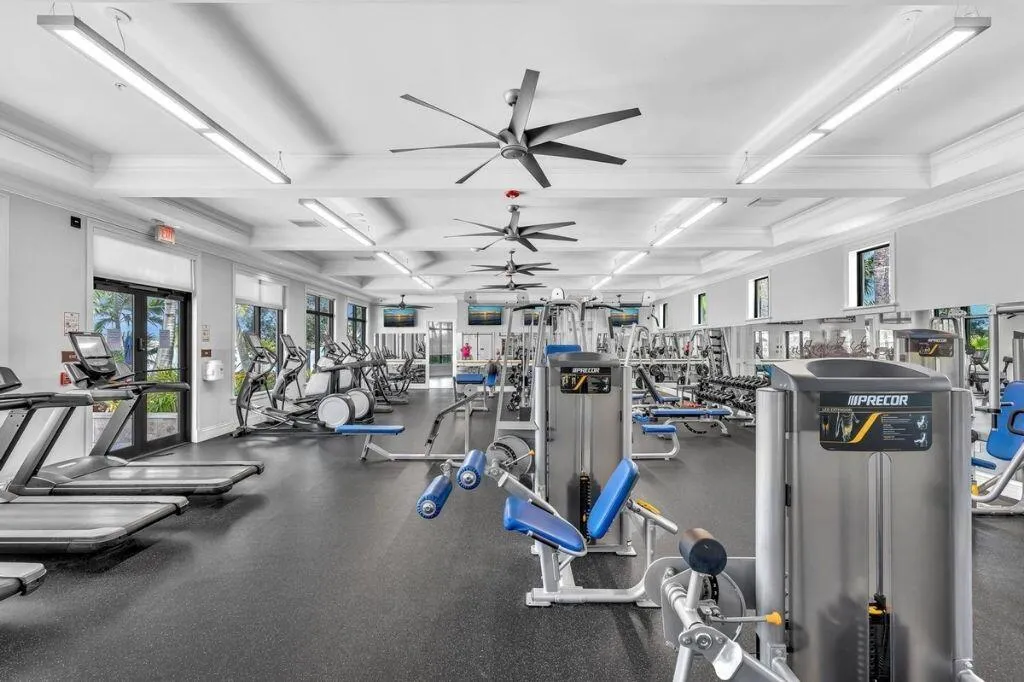 Spacious fitness center featuring various exercise equipment, resilient rubber flooring, overhead lighting, multiple ceiling fans, and large windows