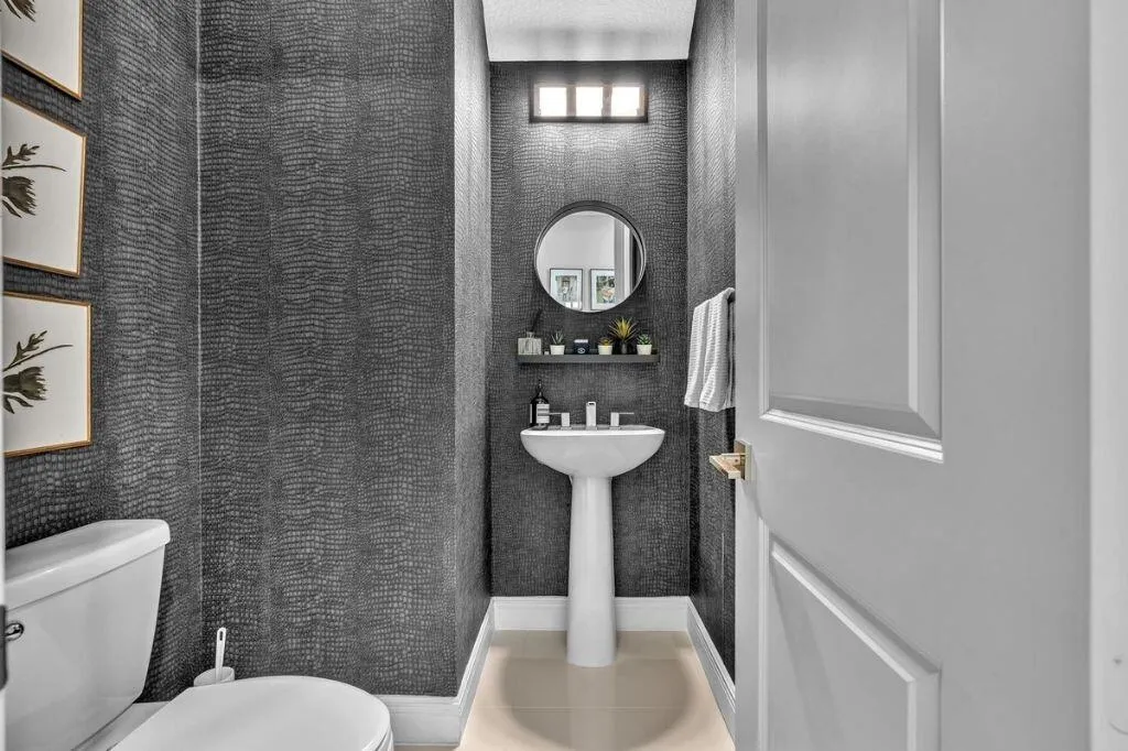 Powder room with textured dark gray wall covering, white pedestal sink, and a circular mirror