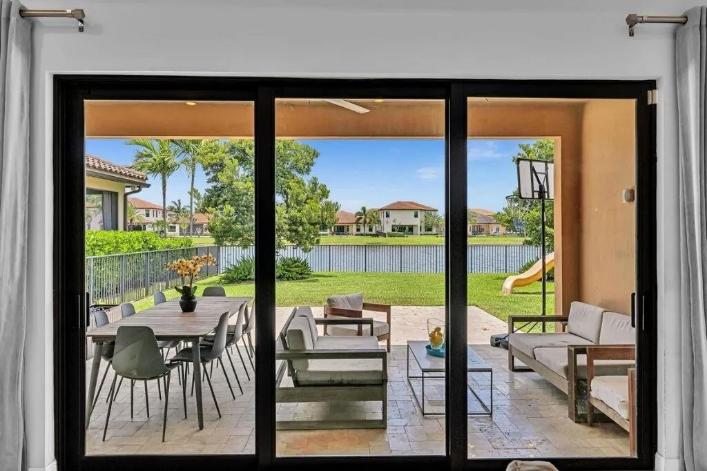 Sliding glass doors opening to a covered patio with tile flooring, featuring outdoor dining and lounge areas, and a manicured lawn overlooking a lake