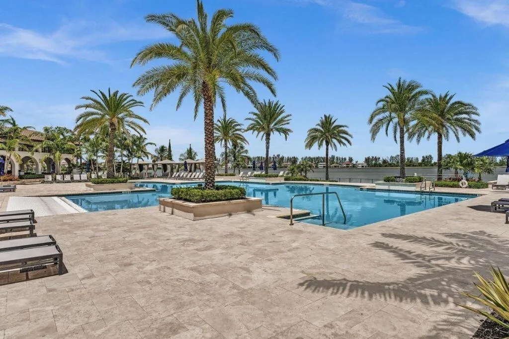 Expansive pool area featuring multiple swimming pools, a stone paver deck, lush landscaping with palm trees, and a waterfront backdrop