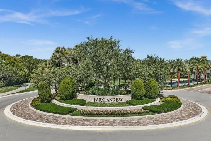 Roundabout entrance featuring a stone-clad "Parkland Bay" sign, lush landscaping, brick paver accents, and mature palm trees
