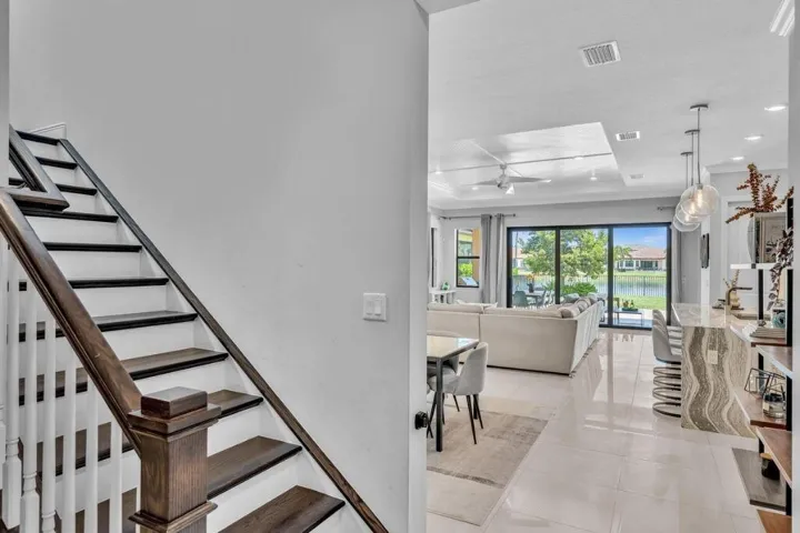 Modern interior featuring a wood-finish staircase with white risers and balusters, a spacious open-concept living area with large format tile flooring, sliding glass doors to outdoor spaces, a kitchen island with a waterfall countertop, and contemporary pendant lighting