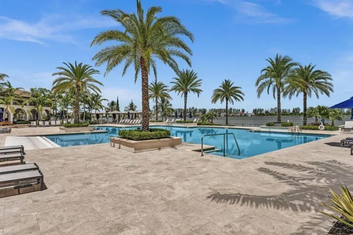 Expansive pool area featuring multiple swimming pools, a stone paver deck, lush landscaping with palm trees, and a waterfront backdrop