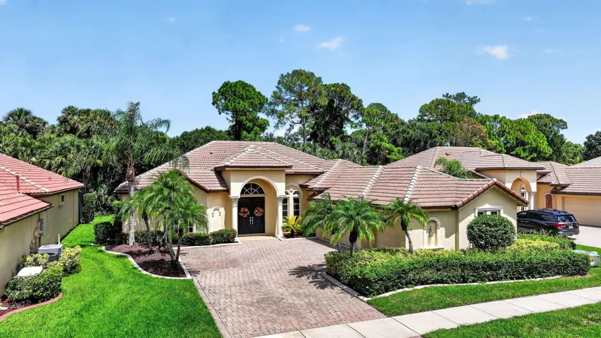 Stucco exterior with arched entry, tile roof, brick paver driveway, mature landscaping, and an attached garage