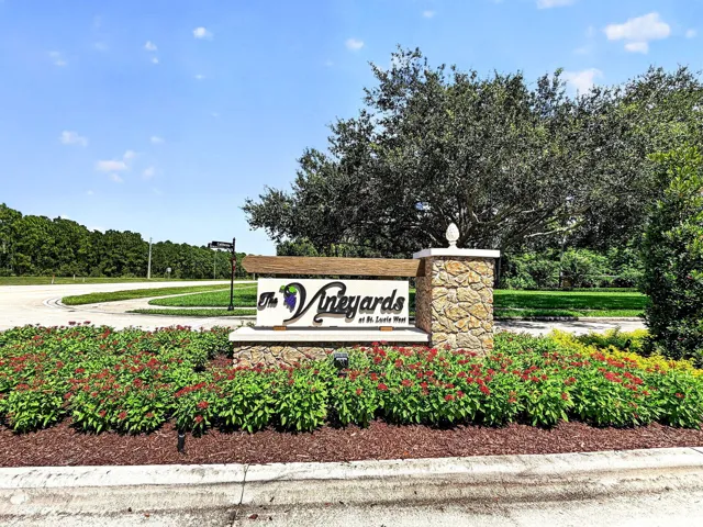 Stone pillar with decorative finial, landscaped entryway with flowering shrubs, and a community sign