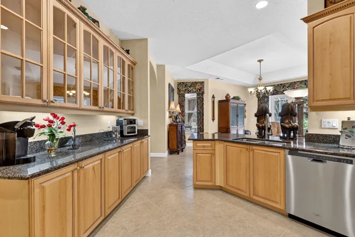 Kitchen featuring light wood cabinetry, glass-front upper cabinets, dark speckled countertops, tile flooring, and stainless steel dishwasher