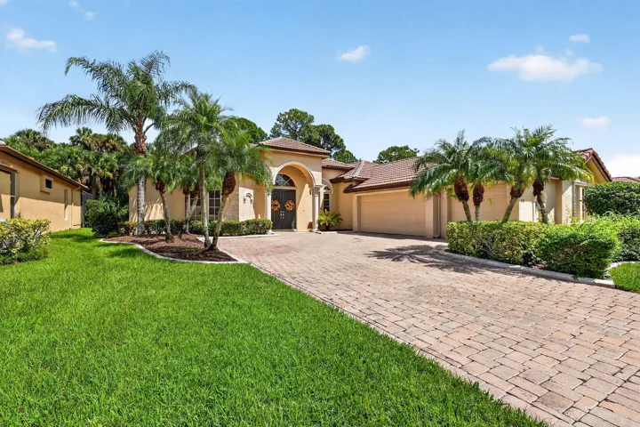 Tan stucco exterior, arched entry, paver driveway, three-car garage, and mature palm trees