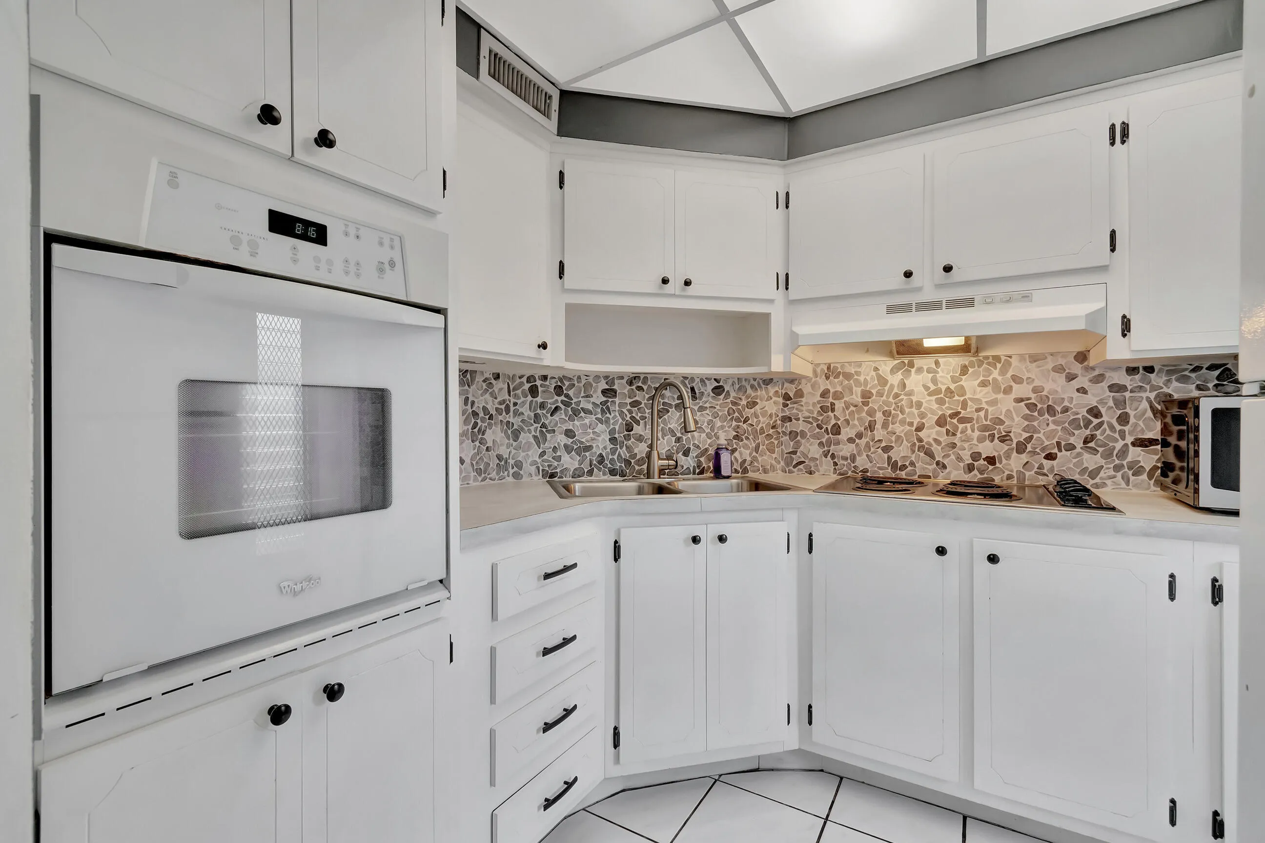 White cabinetry with black hardware, built-in oven, integrated cooktop, pebble-style tile backsplash, and white tile flooring