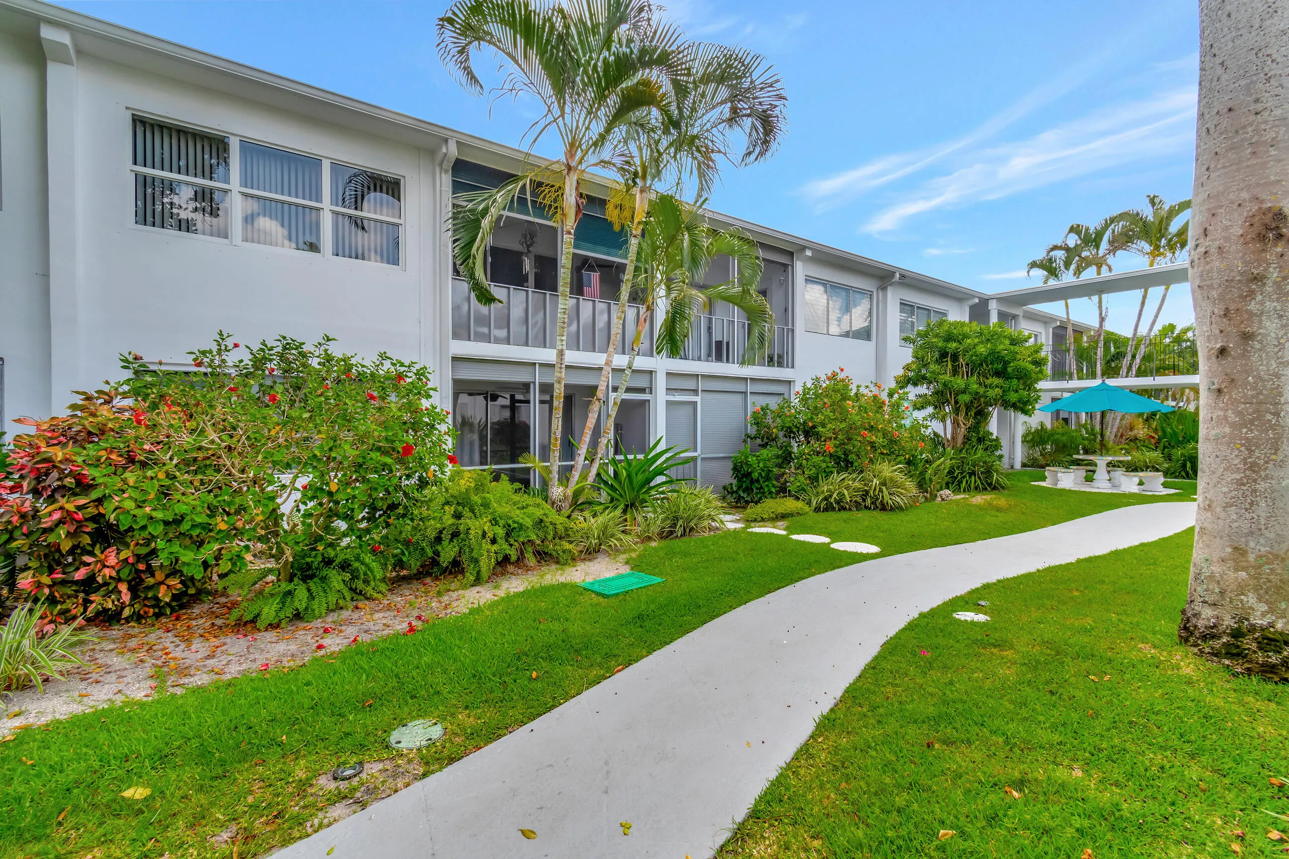Manicured grounds featuring a winding pathway, lush landscaping, mature palm trees, and a shaded outdoor seating area